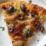 Une part de pizza méditerranéenne aux oignons, anchois et olives noires sur une assiette blanche.